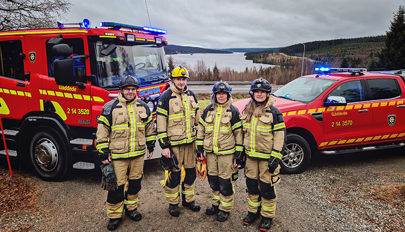Fyra deltidsbrandmän. I bakgrunden vy över Frostviken samt en brandbil och ett räddningsfordon.