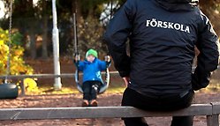 Vuxen i svart jacka med texten Förskola sitter och tittar på ett litet barn som gungar.